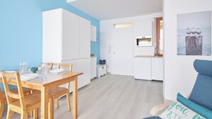 A dining area with a wooden table and chairs, a kitchen in the background, and a blue wall.