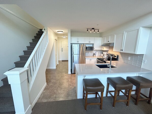 A modern kitchen with white cabinets, a stainless steel refrigerator, a microwave, and a gas stove. There is a wooden island with bar stools. - Penthouse Suite on Golf Course- 308 (Kelowna)