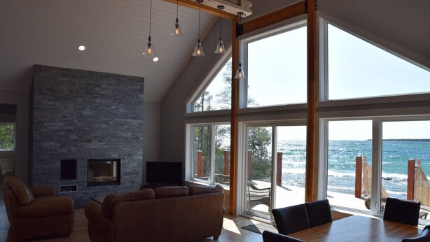 Ein geräumiges Wohnzimmer mit Kamin aus Stein, großen Fenstern mit Meerblick und einem Essbereich mit Holztisch und Stühlen.