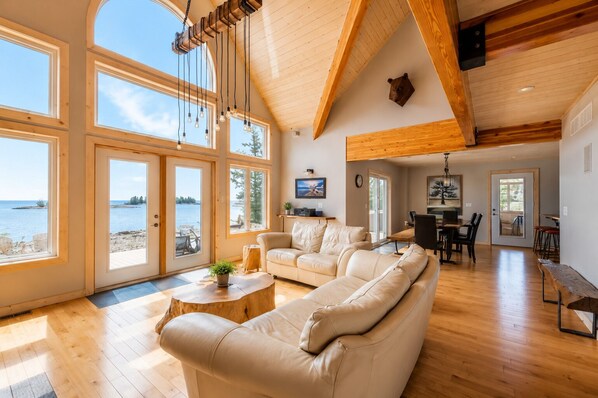 A spacious living room with a large sofa, a coffee table, and a dining area with a table and chairs. There are large windows offering a view of the sea.