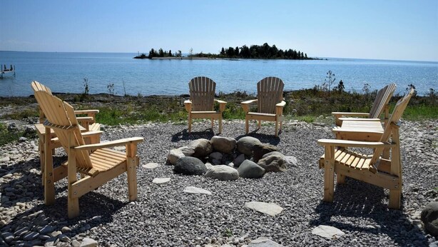 Un paisaje de playa con tres sillas de madera tipo Adirondack dispuestas alrededor de un hogar de piedra, con vista al mar tranquilo y una pequeña isla con árboles.