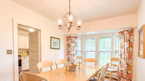 Une salle à manger avec une table en bois, six chaises, un lustre et de grandes fenêtres ornées de rideaux à motifs floraux.