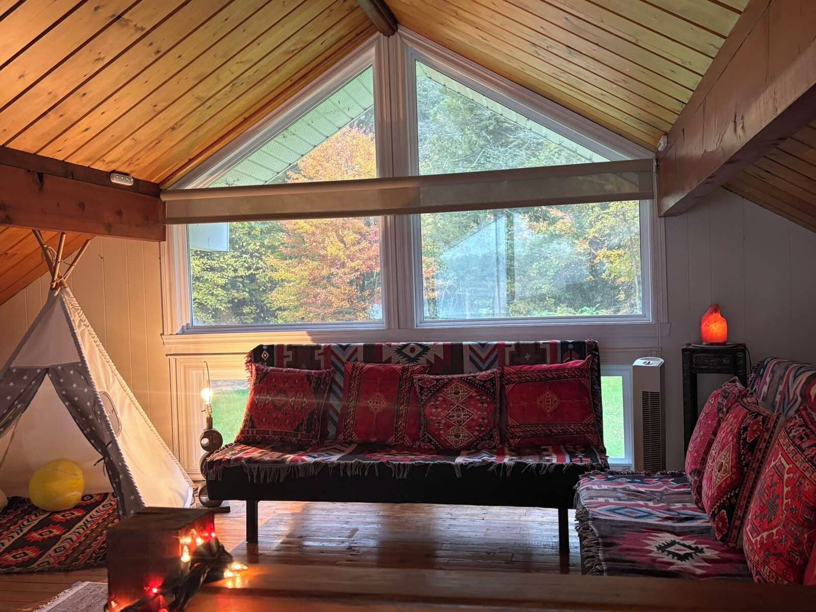 A cozy room with a wooden ceiling, a large window, a sofa with patterned cushions, a small table with a lamp, and a teepee tent.
