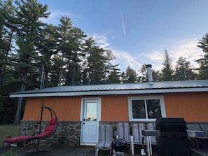 Une cabane avec des fondations en pierre, un toit en métal, une véranda avec des chaises, une chaise suspendue et un barbecue.