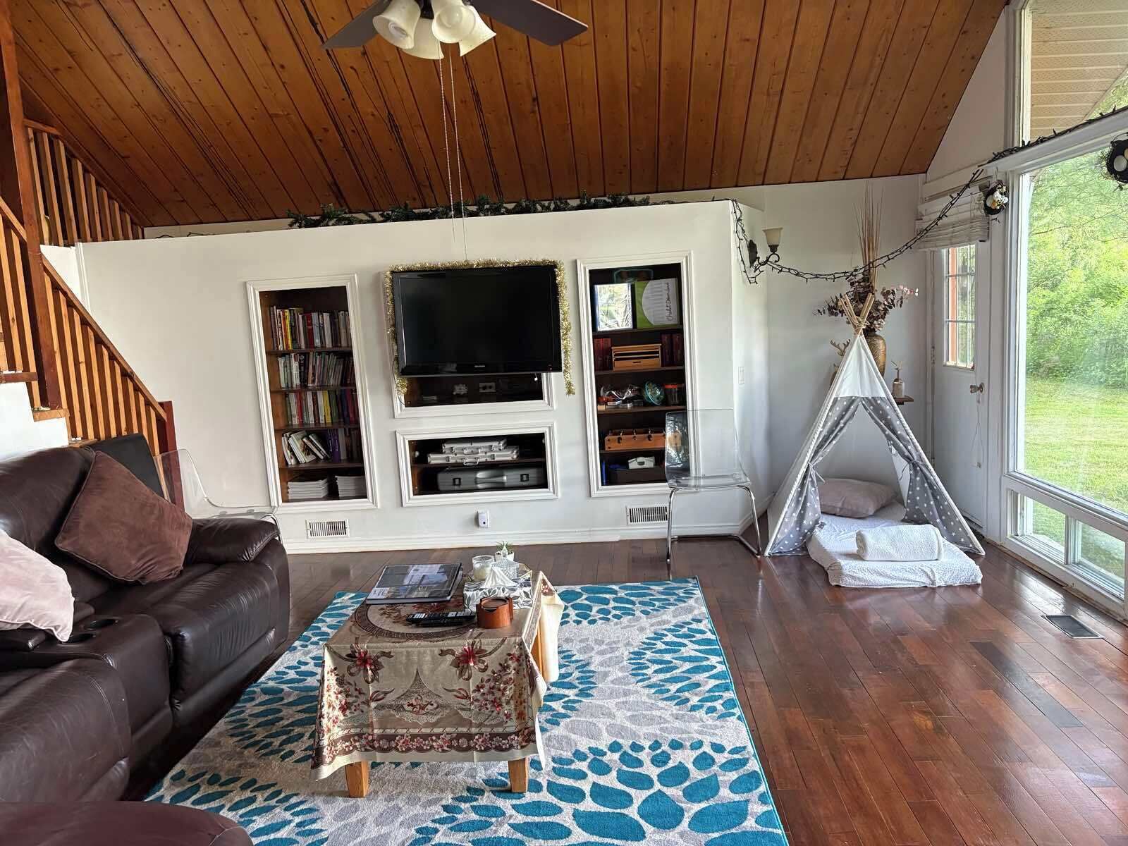 A living room with a wooden ceiling, a TV, bookshelves, a sofa, a coffee table, and a teepee.