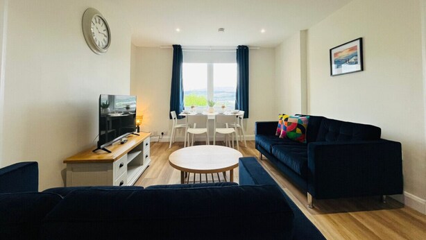A modern living room with a dark blue sofa, a flat-screen TV, a wooden coffee table, and a dining area with a white table and chairs.