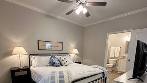 A bedroom with a bed, two bedside tables with lamps, a ceiling fan, a TV on a stand, and a door leading to a bathroom. - The Cottages 3  2BR2BA Dothan Pet Friendly Gorgeous Pool, Pickle Ball court, Gym (Dothan)