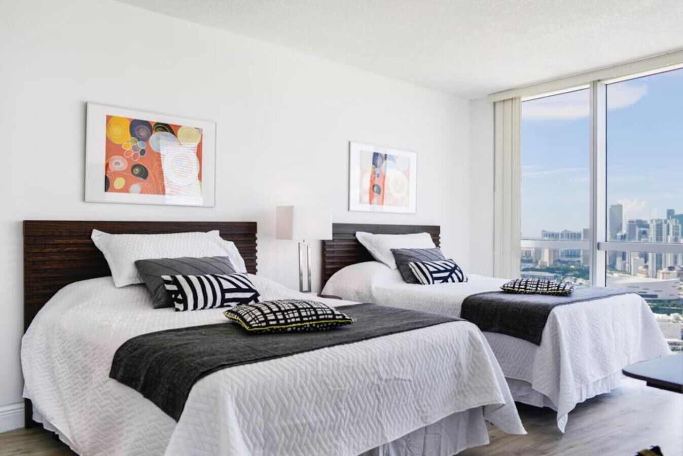 A hotel room with two beds, white bedding, and city views through large windows.