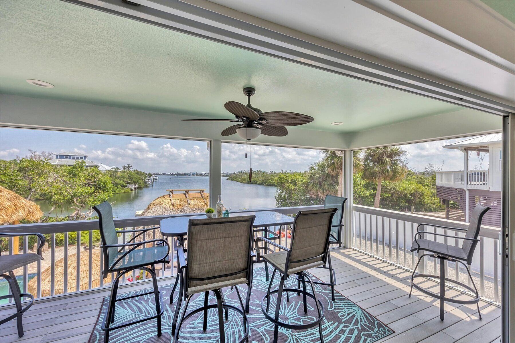 A covered outdoor area with a table and chairs, a ceiling fan, and a view of a body of water and greenery.