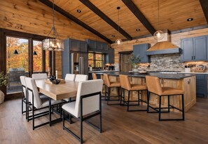 A modern kitchen with wooden beams, a dining area with a large table and chairs, and a view of autumn trees through the window. - Sippin on Sunsets: Ultra Luxe Escape • Epic Views • Hot-Tub • Secluded Comfort (Cherry Log)