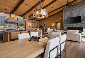 A spacious living area with a large wooden dining table, comfortable seating, a fireplace, and a stone accent wall. - Sippin on Sunsets: Ultra Luxe Escape • Epic Views • Hot-Tub • Secluded Comfort (Cherry Log)