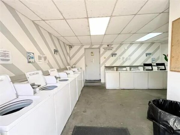 Une buanderie équipée de plusieurs machines à laver et de plusieurs sèche-linge, d’une poubelle et d’une porte donnant sur une autre pièce.