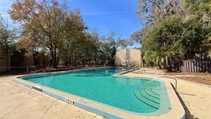 Una piscina rectangular con borde de baldosas, rodeada de arena y árboles.