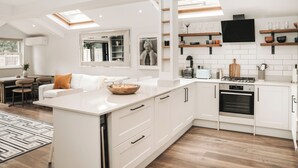 Uma cozinha moderna com armários brancos, uma ilha central e forno embutido. Há um espaço de refeições com um sofá branco e uma mesa de centro.