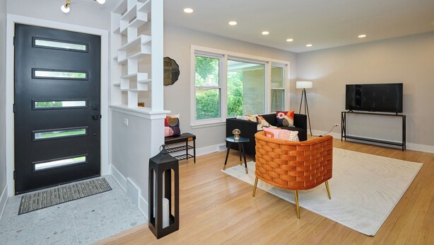 A modern living room with a black door, white shelving, a brick-patterned chair, a black TV stand, and a large window.