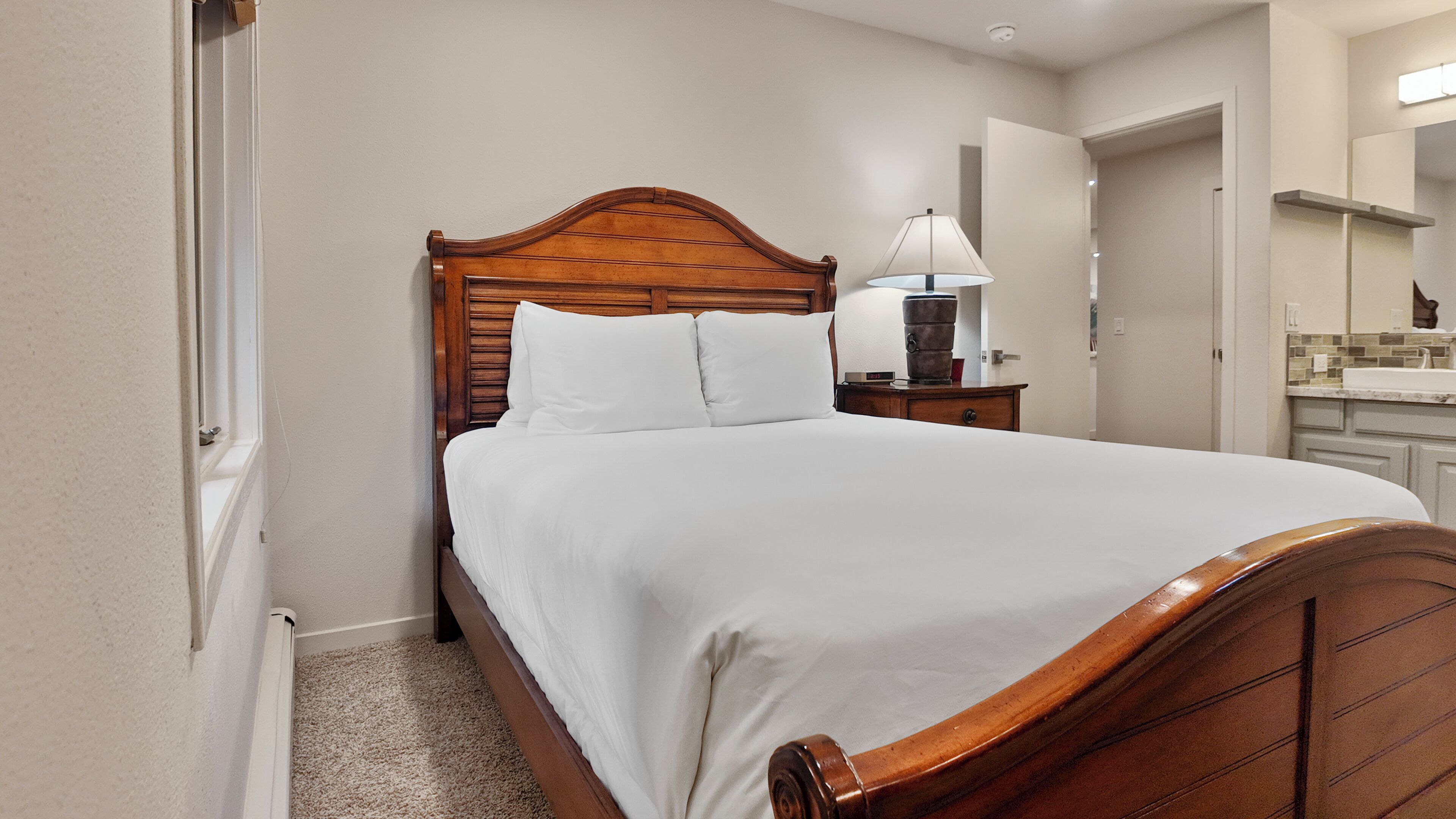 A neatly made bed with a wooden headboard, a bedside table with a lamp, and a bathroom visible in the background.
