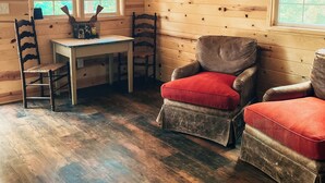 A rustic cabin interior with wooden walls, a wooden ceiling, a dining table, two armchairs, and a window with a view of greenery. - The Cabins at Bent Pine Ranch ~ Cabin #1 (Locust Grove)