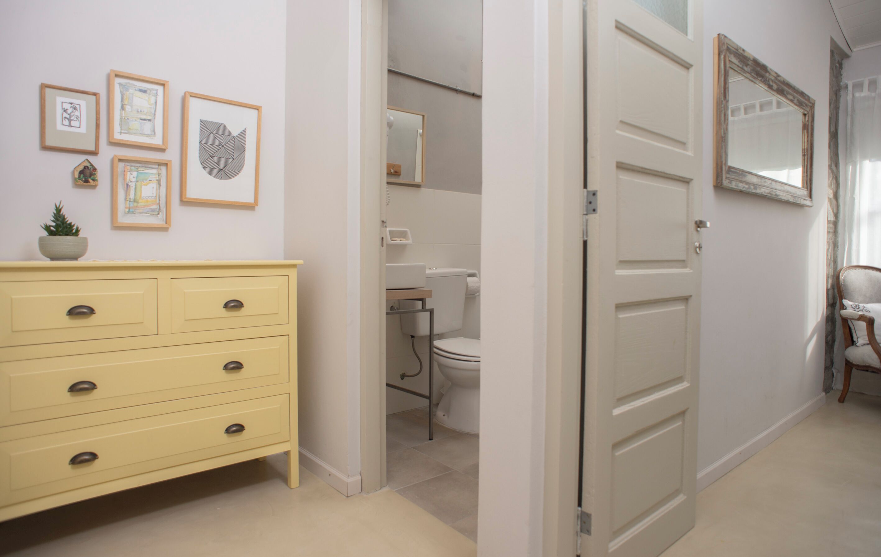Un couloir avec une commode jaune, des œuvres d’art encadrées et une porte donnant sur une salle de bain équipée de toilettes et d’un lavabo.