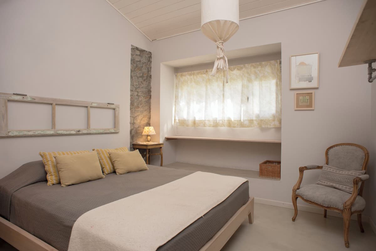 Ein Schlafzimmer mit einem Bett, einem Sessel, einem Fenster mit Vorhängen, einer Lampe und einem an der Wand befestigten Regal.