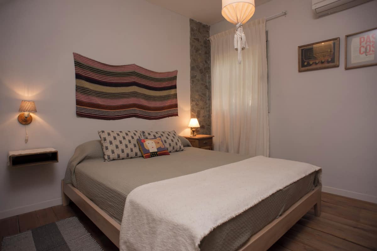 Ein Schlafzimmer mit einem Bett, einer Wandlampe, einem Nachttisch, einem Teppich und einer großen, gestreiften Wandtextilie.