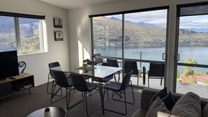Ein modernes Wohnzimmer mit großem Fenster, das auf einen See und Berge blickt. Es gibt einen Esstisch mit Stühlen, eine Couch und einen Fernseher auf einem Schrank.