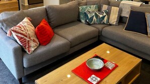 Un salon moderne avec un canapé d’angle gris, une table basse en bois et un coin cuisine équipé d’un réfrigérateur et de tabourets de bar.