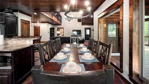 A dining room with a long wooden table set for six, flanked by dark wooden cabinets and a kitchen area with a built-in microwave.