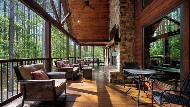 A spacious wooden deck with wicker furniture, a stone fireplace, and large windows offering forest views.