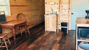 A wooden cabin interior with a dining table, chairs, a kitchen area with a sink and stove, and a ladder leading to a lofted sleeping area.