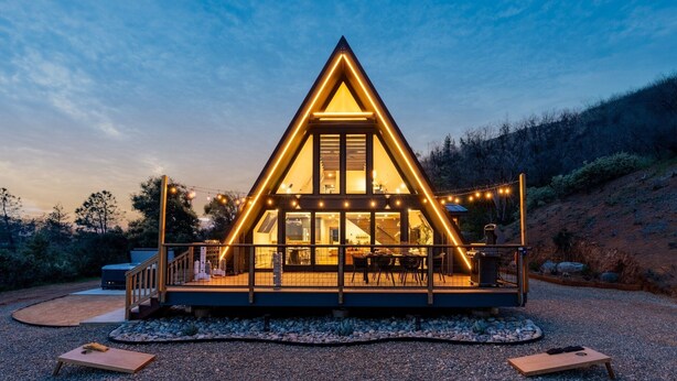 A triangular wooden cabin with a balcony, string lights, and a gravel driveway.