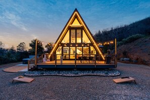 A triangular wooden cabin with a balcony, string lights, and a gravel driveway.