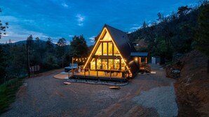 A modern, triangular-shaped cabin with large windows, a hot tub, and a deck area, surrounded by a forested landscape.