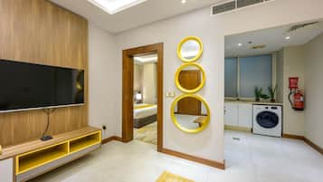 Una habitación de hotel moderna con televisor de pantalla plana, mueble de entretenimiento de madera, pared espejada, lavadora y pequeña cocina.