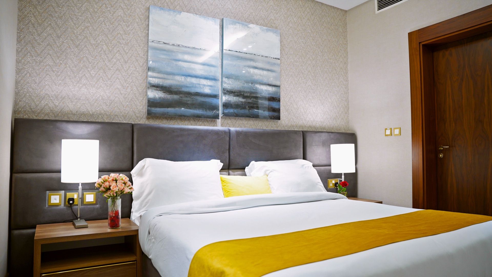 A hotel room with a large bed, two bedside lamps, a nightstand with a vase of flowers, and a wooden door.