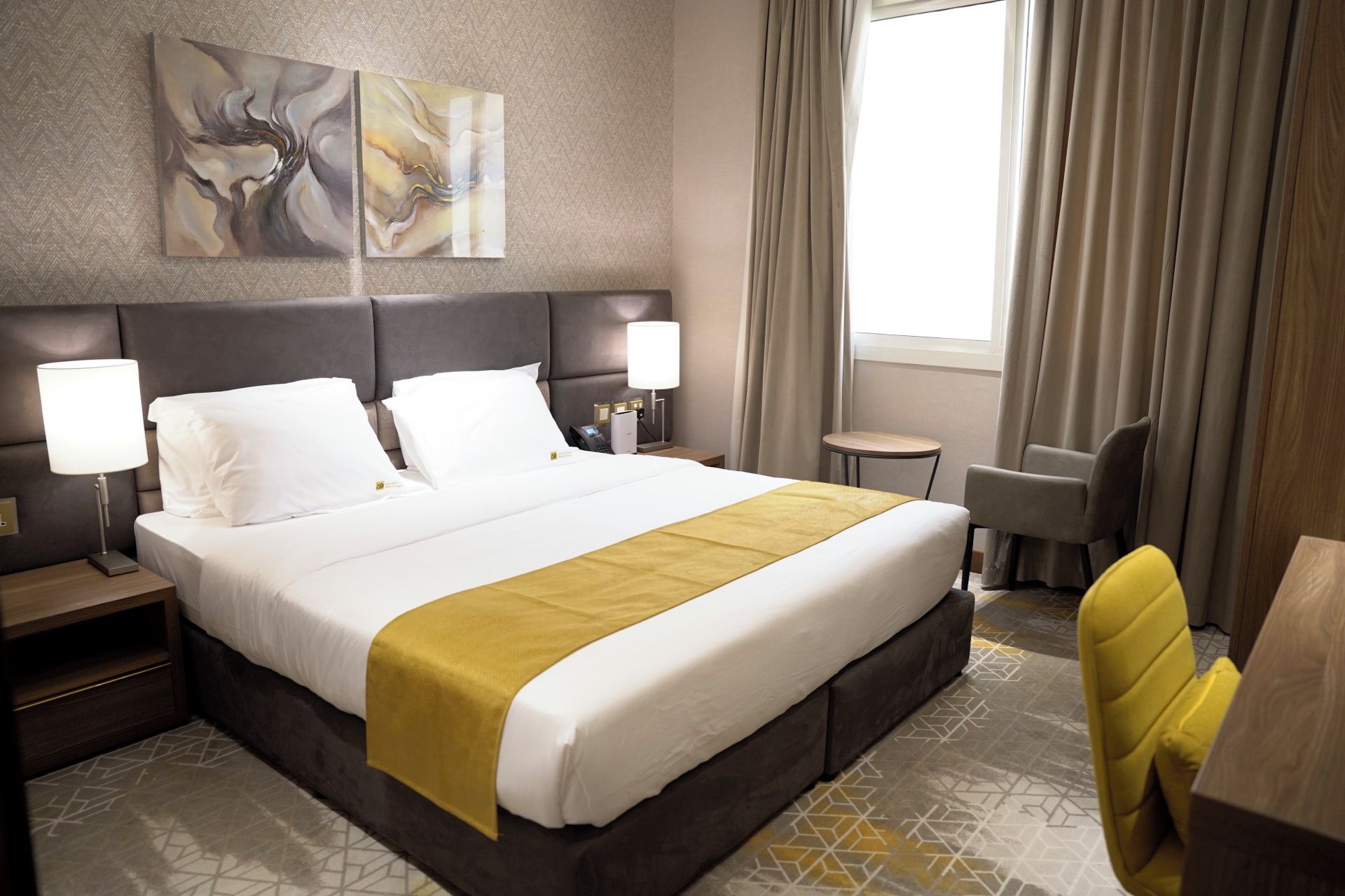 A hotel room with a large bed, two bedside tables, a desk, a chair, and a window with curtains.