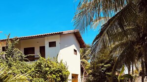 Un edificio blanco con tejado de baldosas, rodeado de abundante vegetación y una piscina.