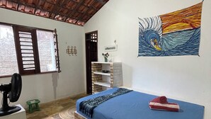 Una habitación con una cama, un ventilador, un taburete pequeño, un estante y un cuadro en la pared.