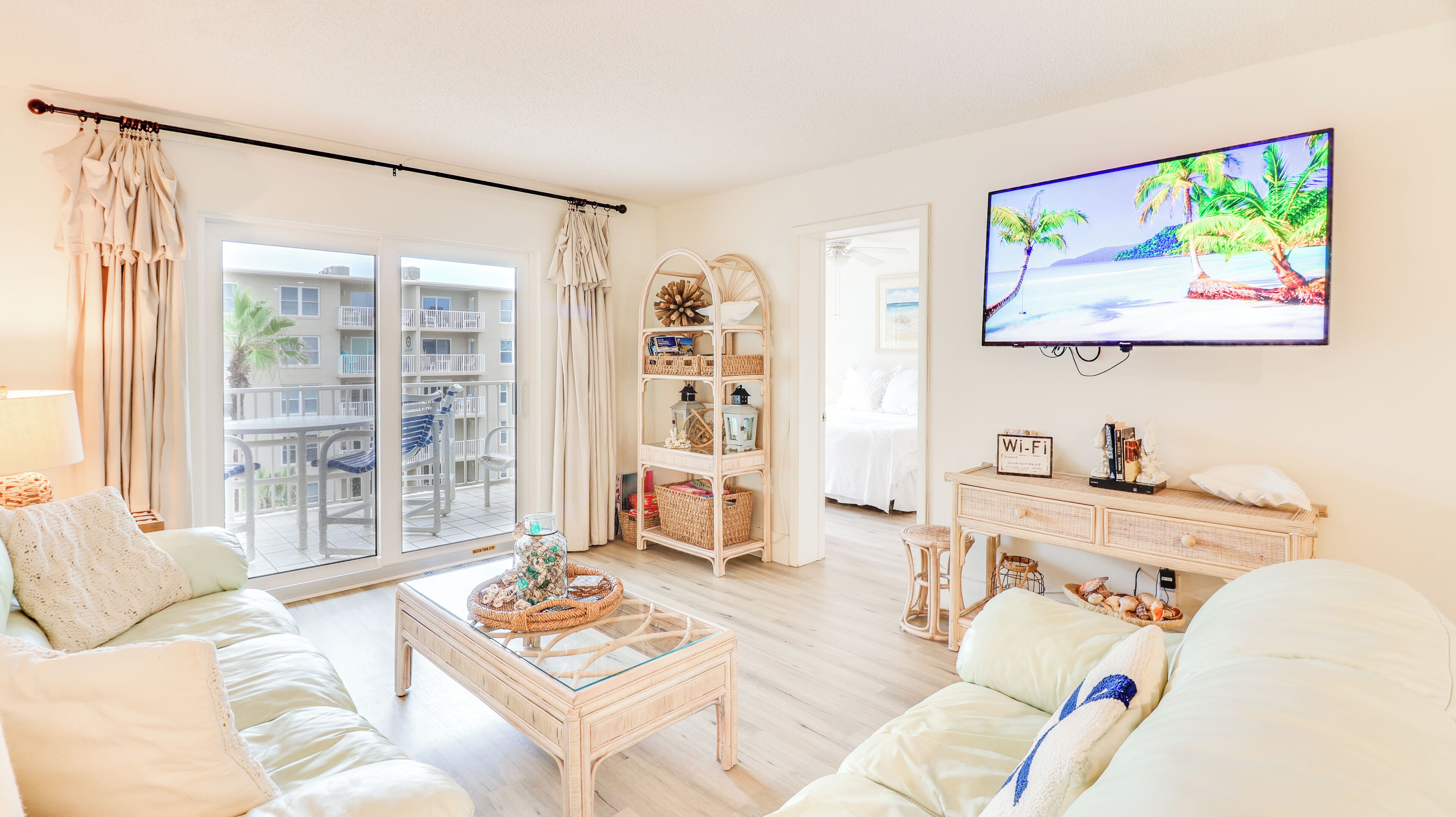 A living room with a sofa, coffee table, and TV. There is a balcony with a view of other buildings and a palm tree.