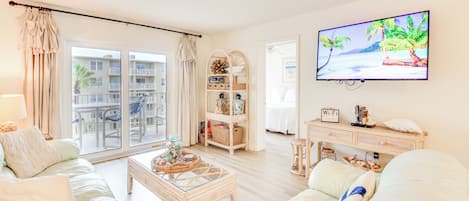 A living room with a sofa, coffee table, and TV. There is a balcony with a view of other buildings and a palm tree.
