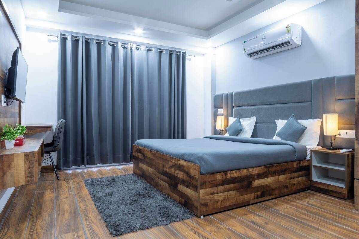 A modern bedroom with a wooden bed frame, a desk with a chair, a TV mounted on the wall, and a large window with curtains.