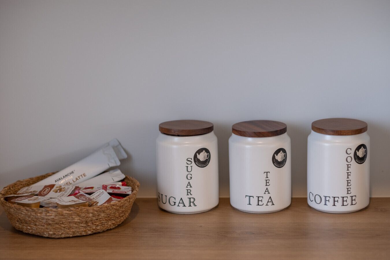 Three white containers with brown lids labeled 'SUGAR,' 'TEA,' and 'COFFEE' on a wooden surface.