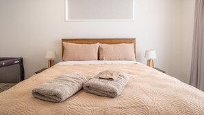 A neatly made bed with a beige quilt, two pillows, and two folded towels on a wooden headboard. There are two bedside lamps and a window with a blind.