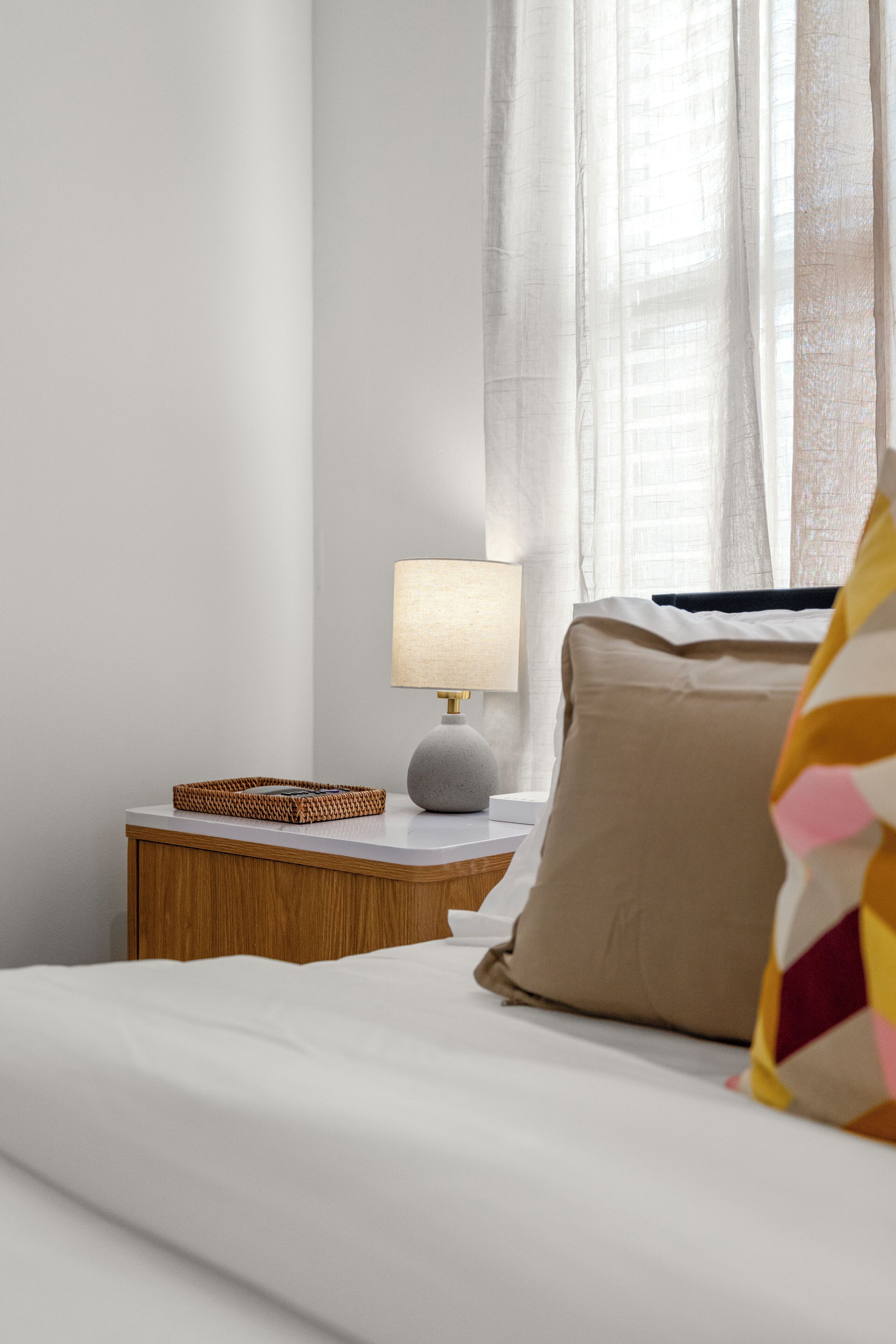 A neatly made bed with a white comforter, a wooden nightstand with a lamp, and a woven basket.
