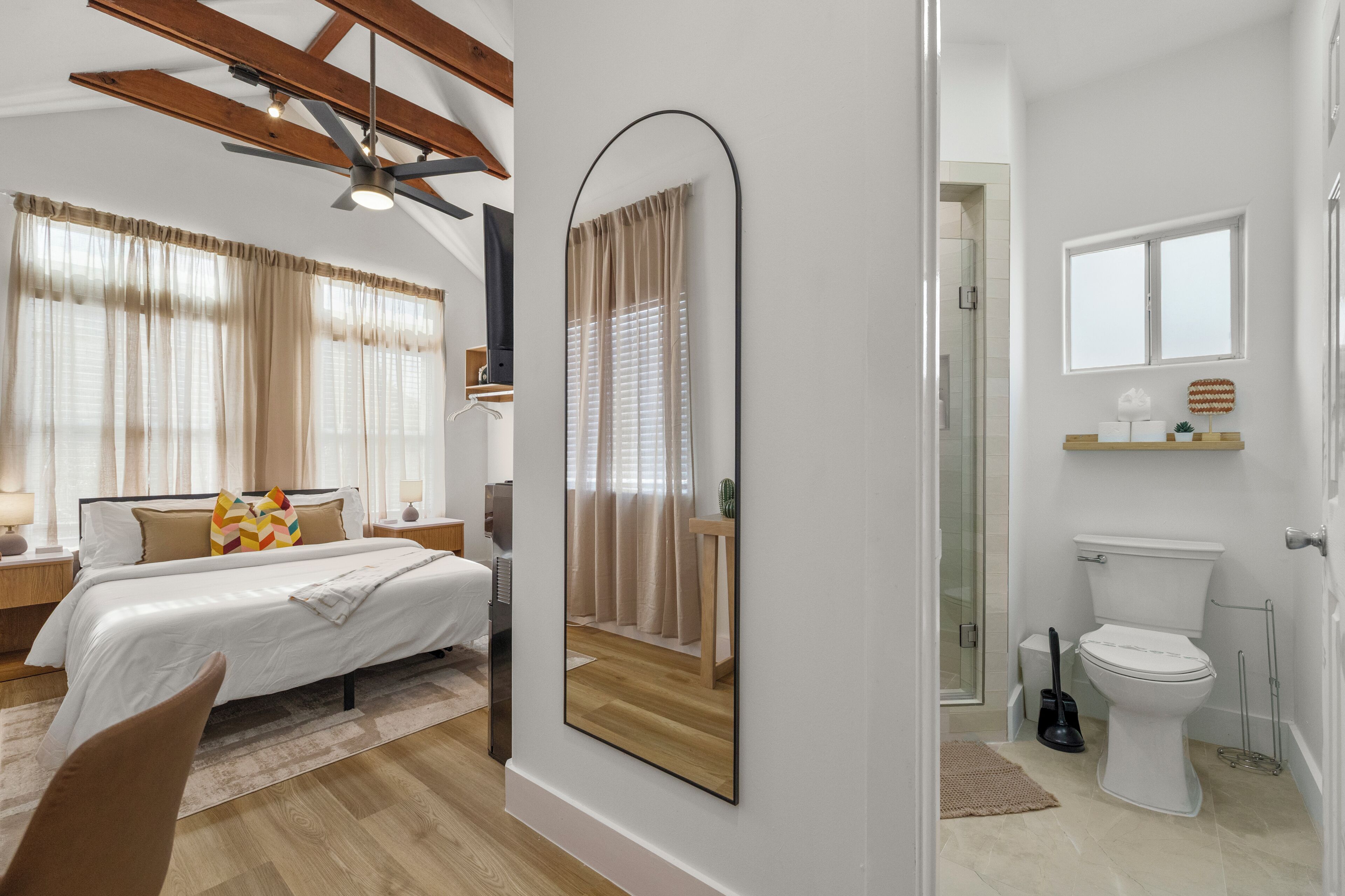 A modern bedroom with a large bed, a bedside table, and a nightstand. There is a bathroom with a toilet, a sink, and a mirror. The room has wooden flooring and a ceiling fan.