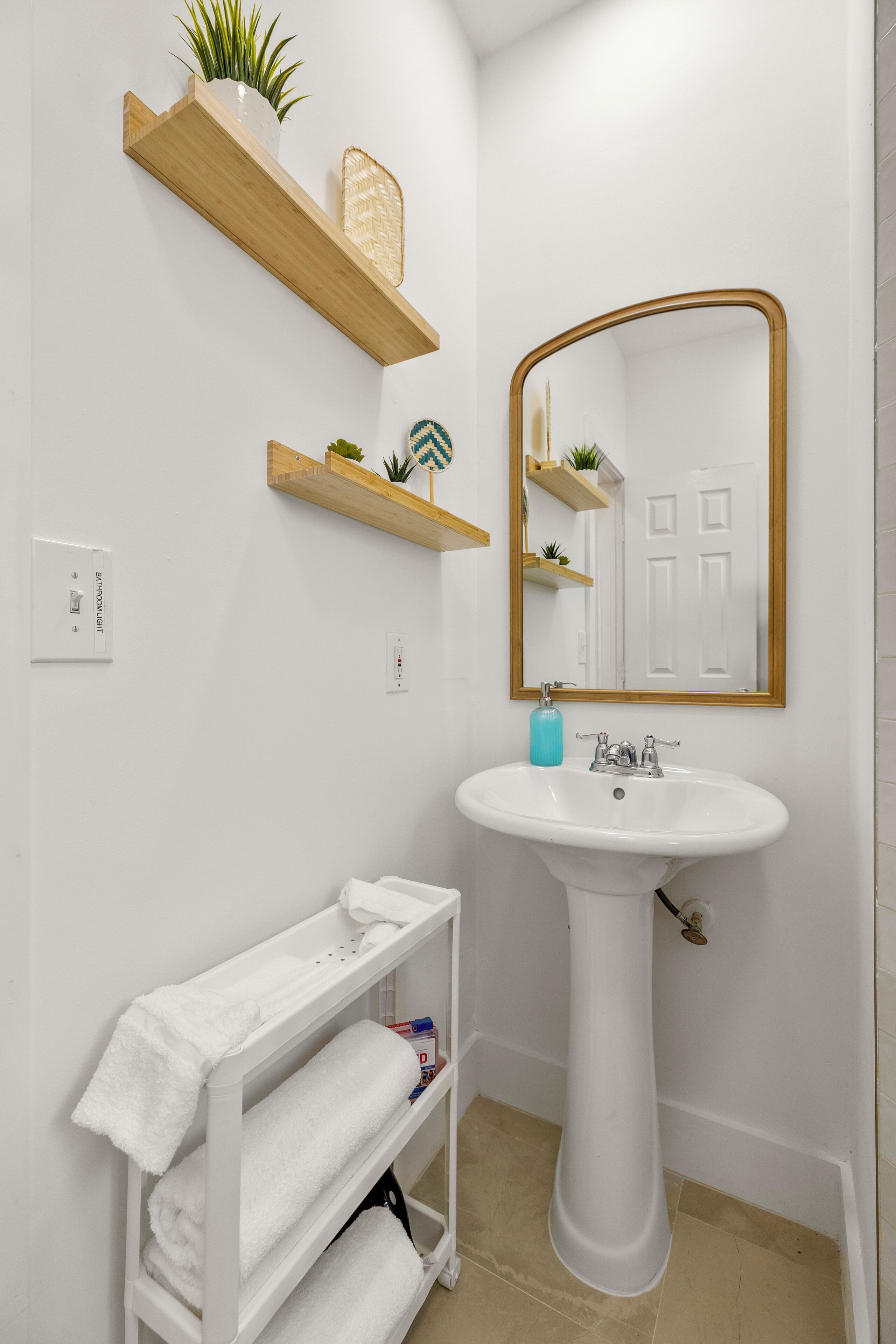 A bathroom with a pedestal sink, wooden shelves with plants, a towel rack, and a mirror with a gold frame.