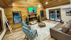 A cozy living room with wooden walls, a fireplace, a television, and a large window. - "Pike's Place" New Cabin in Daisy Ark. (Kirby)