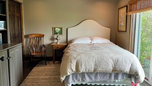 A bedroom with a bed, a chair, a nightstand, and a window with a view of greenery. - Bozeman Farmhouse Cottage (Bozeman)