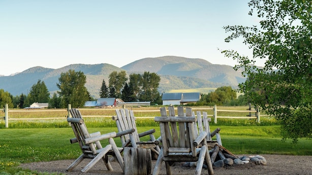 A rustic outdoor seating area with wooden chairs and a fire pit, set against a backdrop of mountains and a clear sky. - Bozeman Farmhouse Cottage (Bozeman)