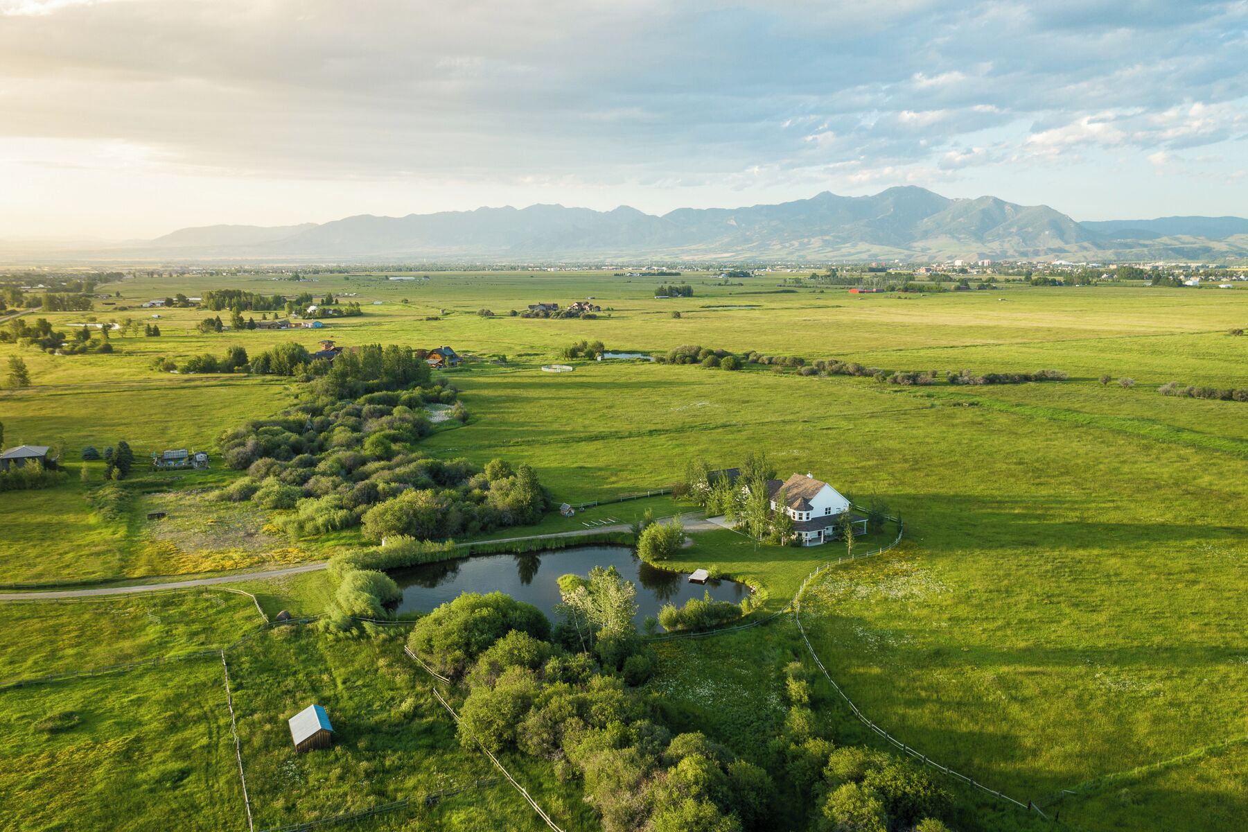 A rural landscape with a small house near a pond, surrounded by fields and mountains in the background.