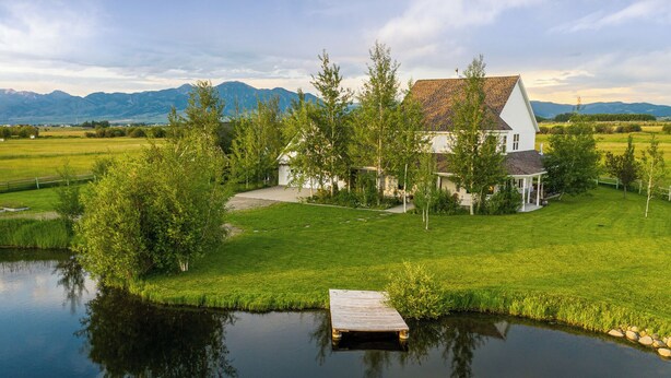 Ein Haus mit Teich davor, umgeben von Grünflächen und Bergen im Hintergrund.